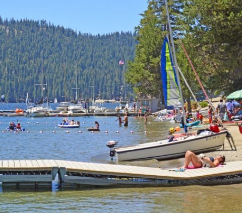 Beach scene: Lake with docked boats, people swimming and sunbathing, sailboat on shore, forest backdrop.