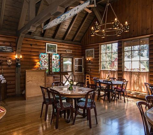 Interior of a rustic lodge with wood paneling, tables, chairs, and a chandelier; light streams in from large windows.