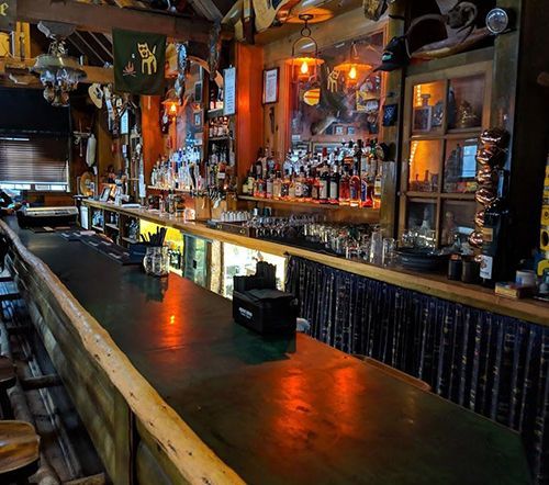 Wooden bar in a dimly lit bar, lined with bottles and glasses. Rustic decor with hanging ornaments.