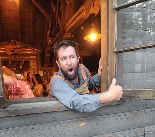Man leans out of a rustic window, giving a thumbs up. He has a surprised expression. A lit lamp hangs above him.