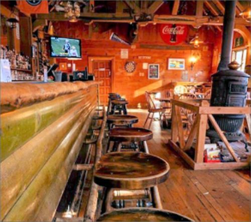 Wooden bar interior with stools, tables, and a wood-burning stove. A TV displays a game. Warm, rustic ambiance.