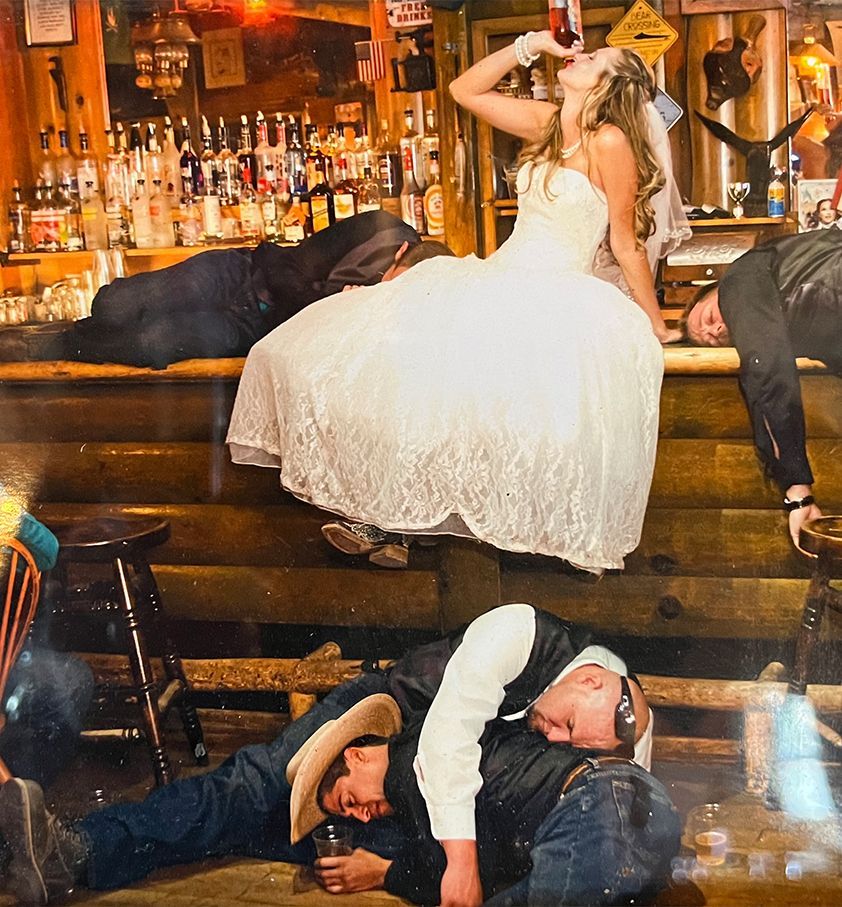 Bride in wedding dress drinking at bar; several people passed out nearby.