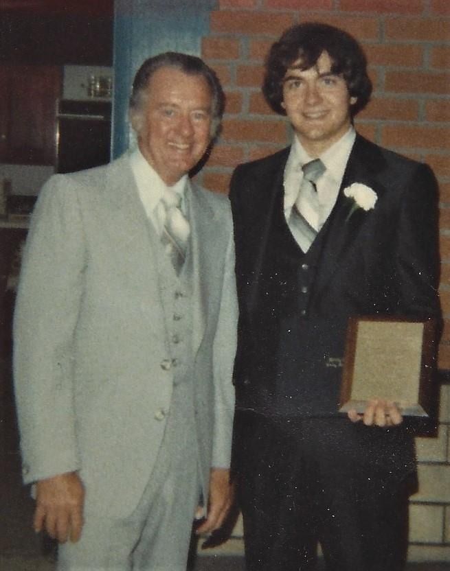 Two men in suits posing indoors; one holds a plaque or award.