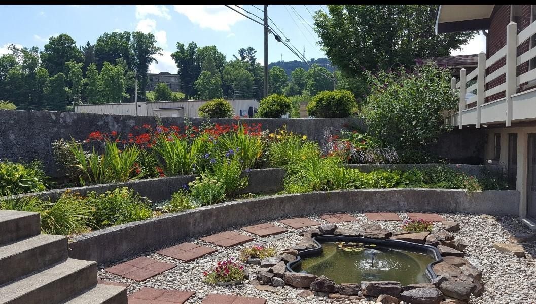 Garden courtyard with a small pond, stone path, and lush green and red plants beside a building.