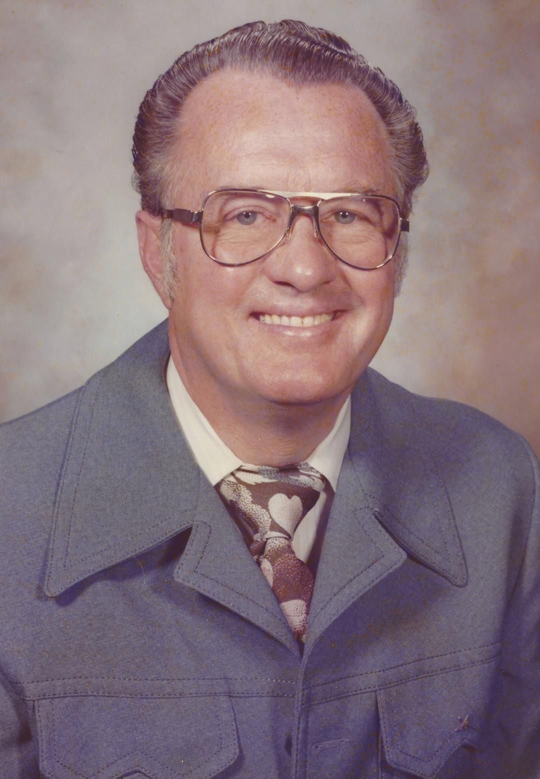 Smiling man in a gray suit, patterned tie, and glasses against a soft studio backdrop