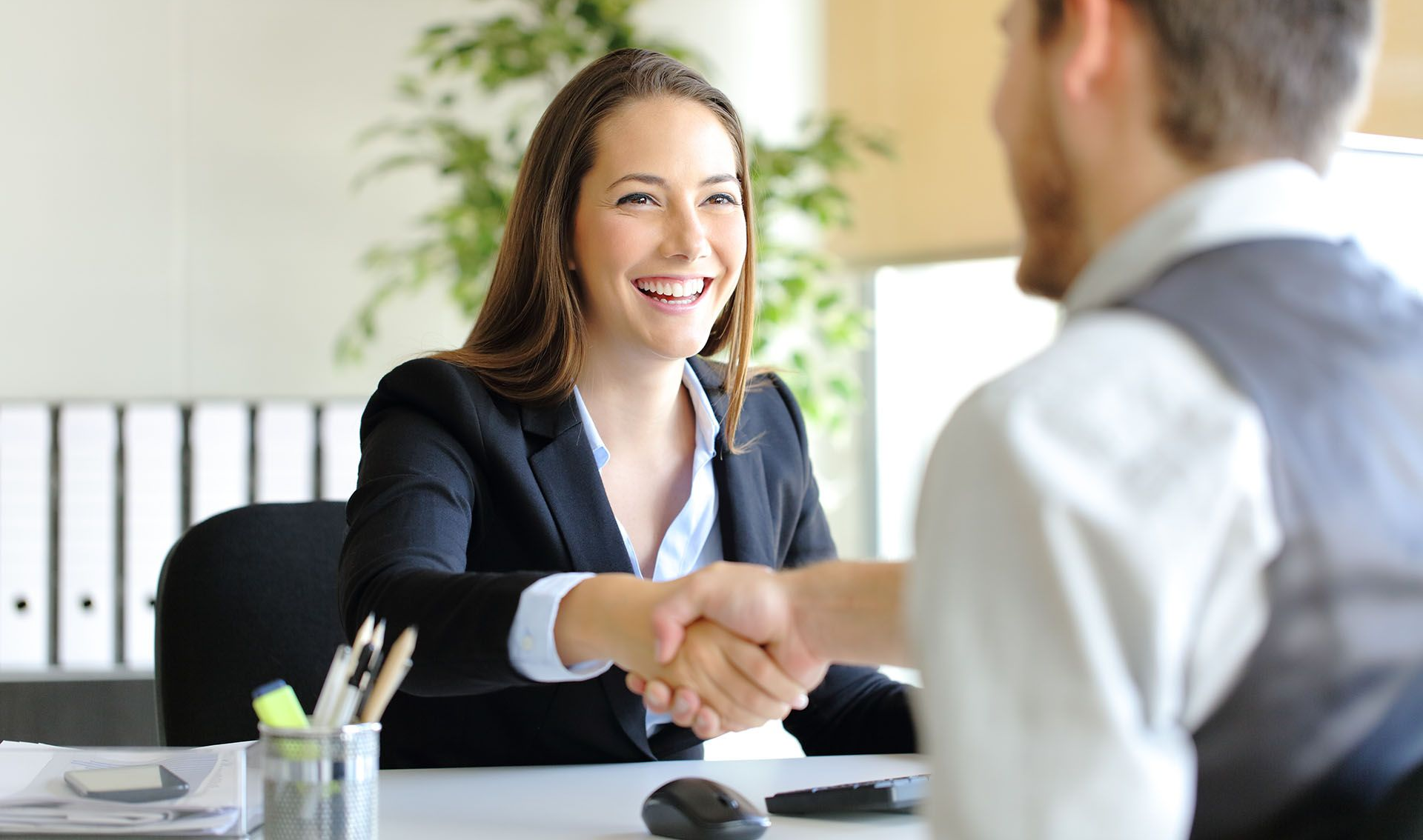 Two professionals shaking hands in an office, smiling across a desk.