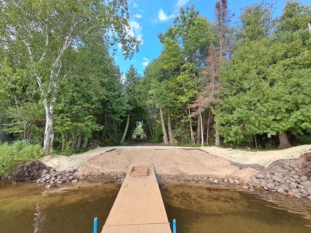 Wooden dock leading to a sandy shoreline, bordered by rocky banks and dense green trees under a blue sky