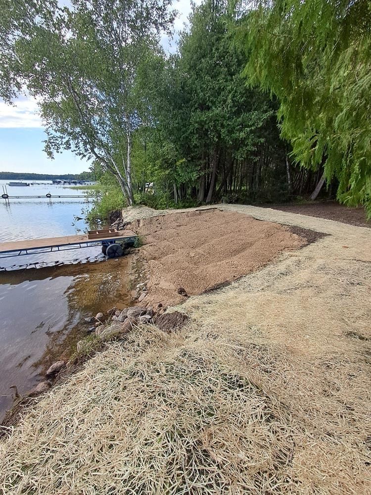 Shoreline path beside a calm lake, with trees, a small dock, and sandy bank.
