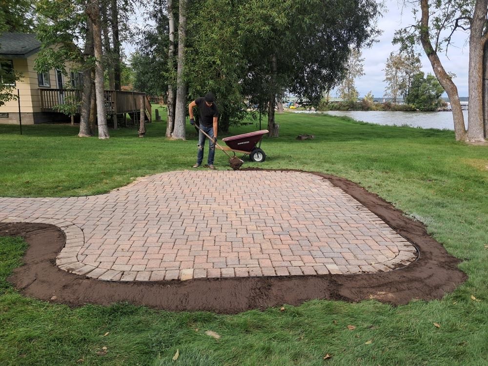 Patio pavers being installed in a grassy yard by a person with a wheelbarrow near trees and a lake