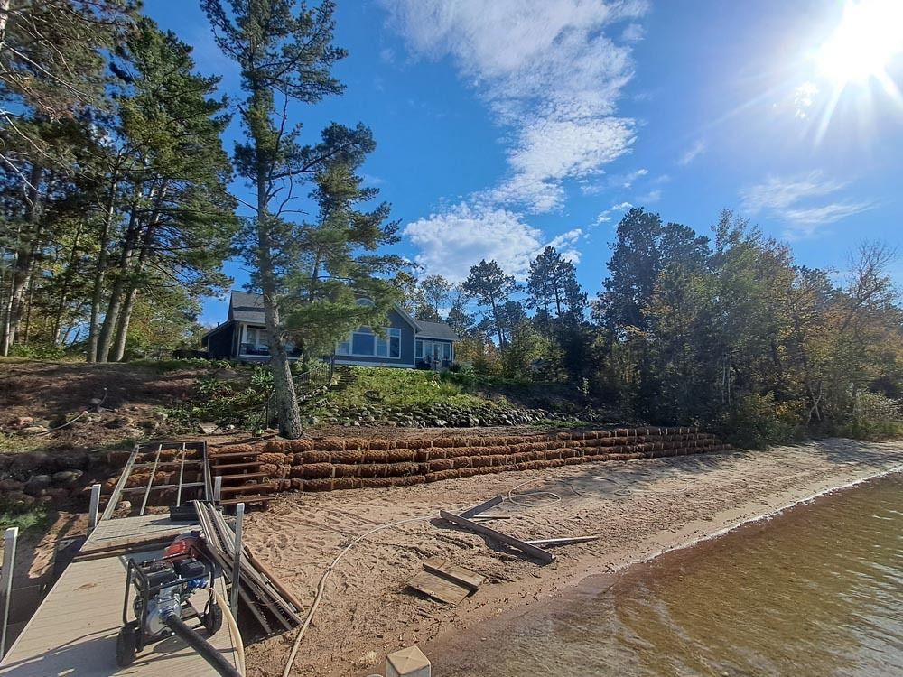Dock and rocky shoreline beside a blue house and pine trees under a bright sunny sky.