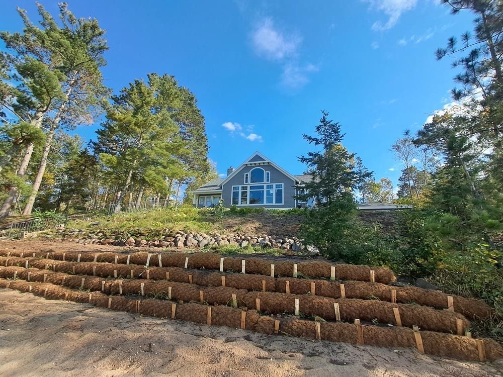 Blue house on a hill with trees, stacked log retaining wall, and bright blue sky