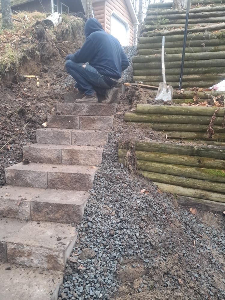 Person crouching beside a gravel path and concrete steps near stacked logs outdoors