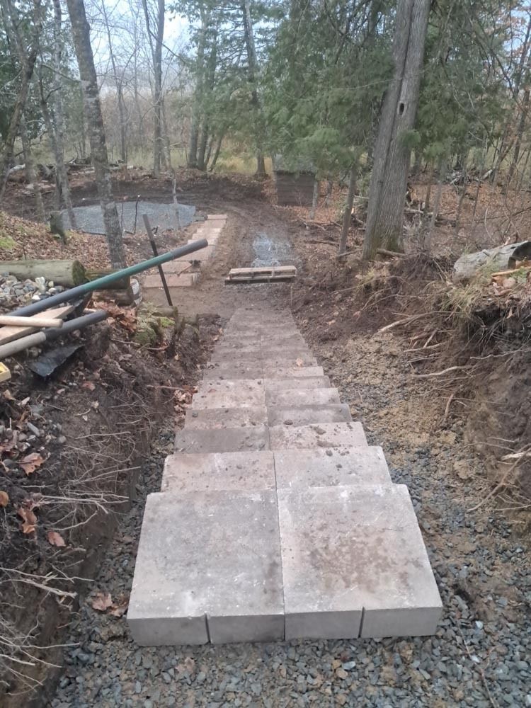 Stone steps descend through a wooded, muddy slope to a small path with a railing on the left.