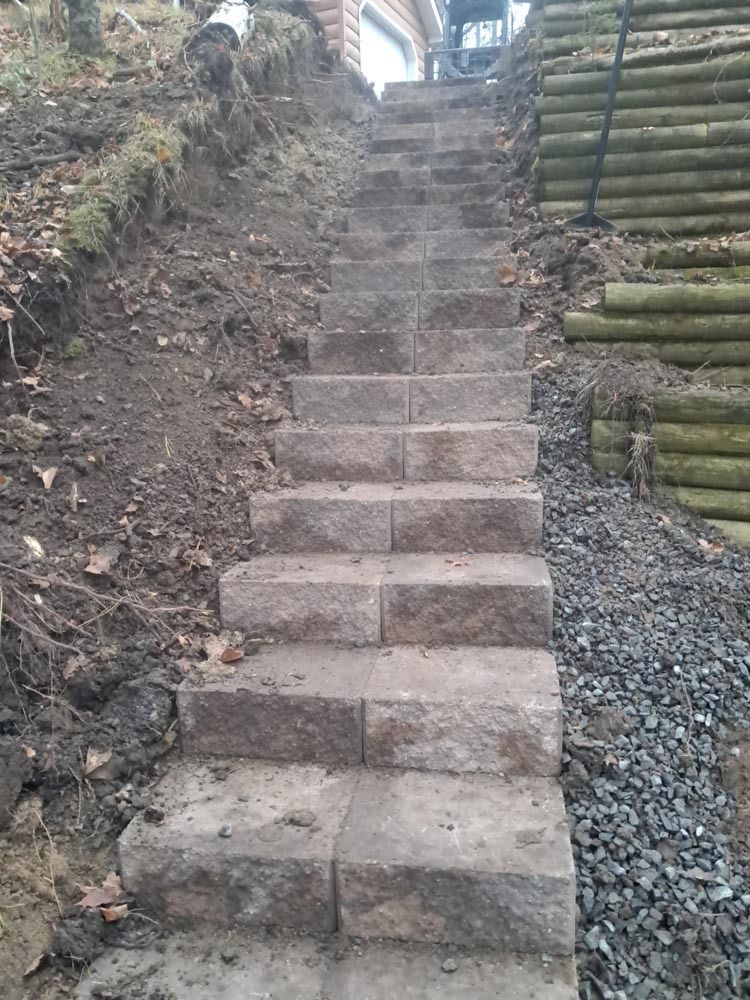 Narrow outdoor stone steps climbing a steep dirt slope with gravel and stacked logs on the side