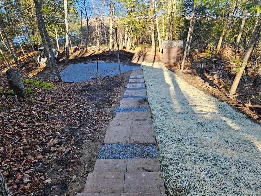 Stone path beside frost-covered lawn in a wooded backyard, with a small frozen pond nearby