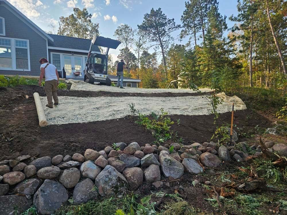 Landscape retaining wall under construction beside a house, with workers and stone border in a wooded yard.