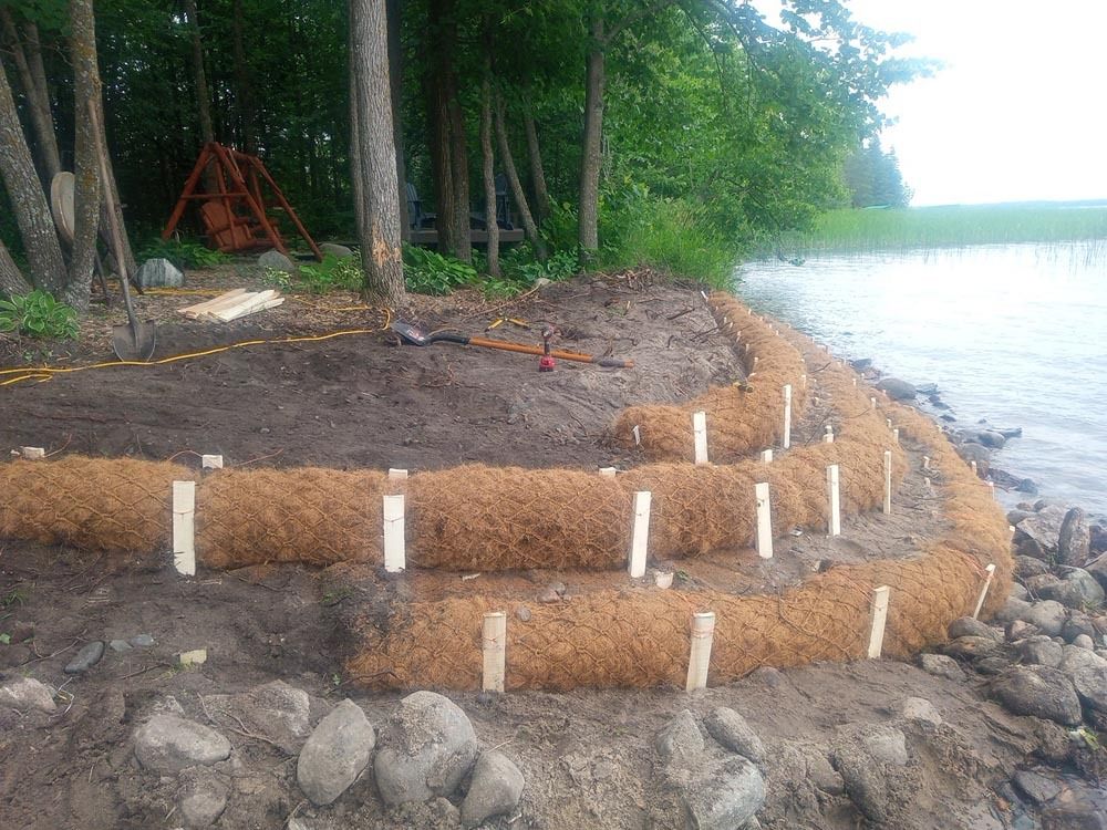 Coastal shoreline erosion control with layered sandbags and white posts beside a wooded lake shore