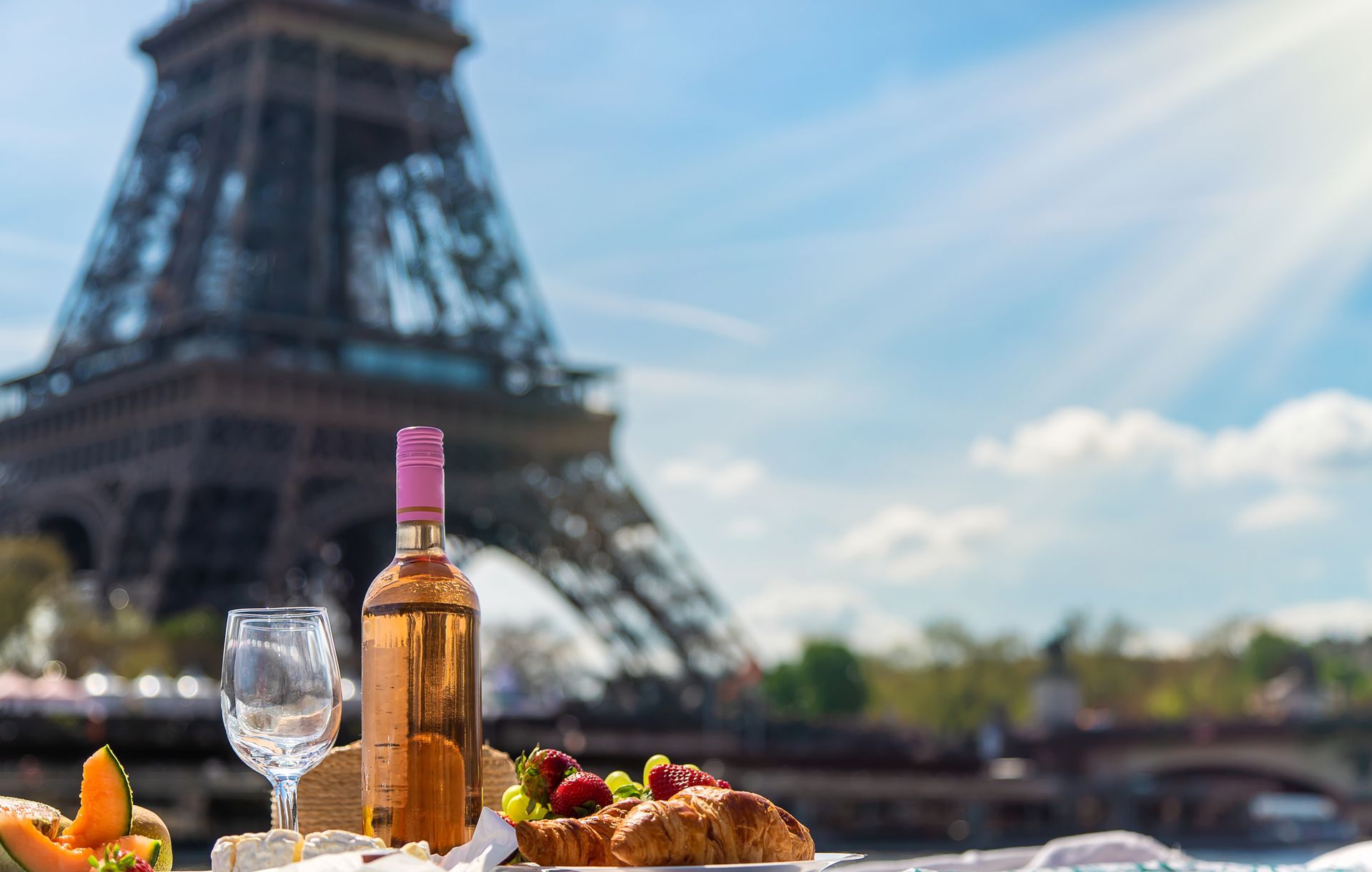 A bottle of wine , a croissant , and two glasses are on a table in front of the eiffel tower