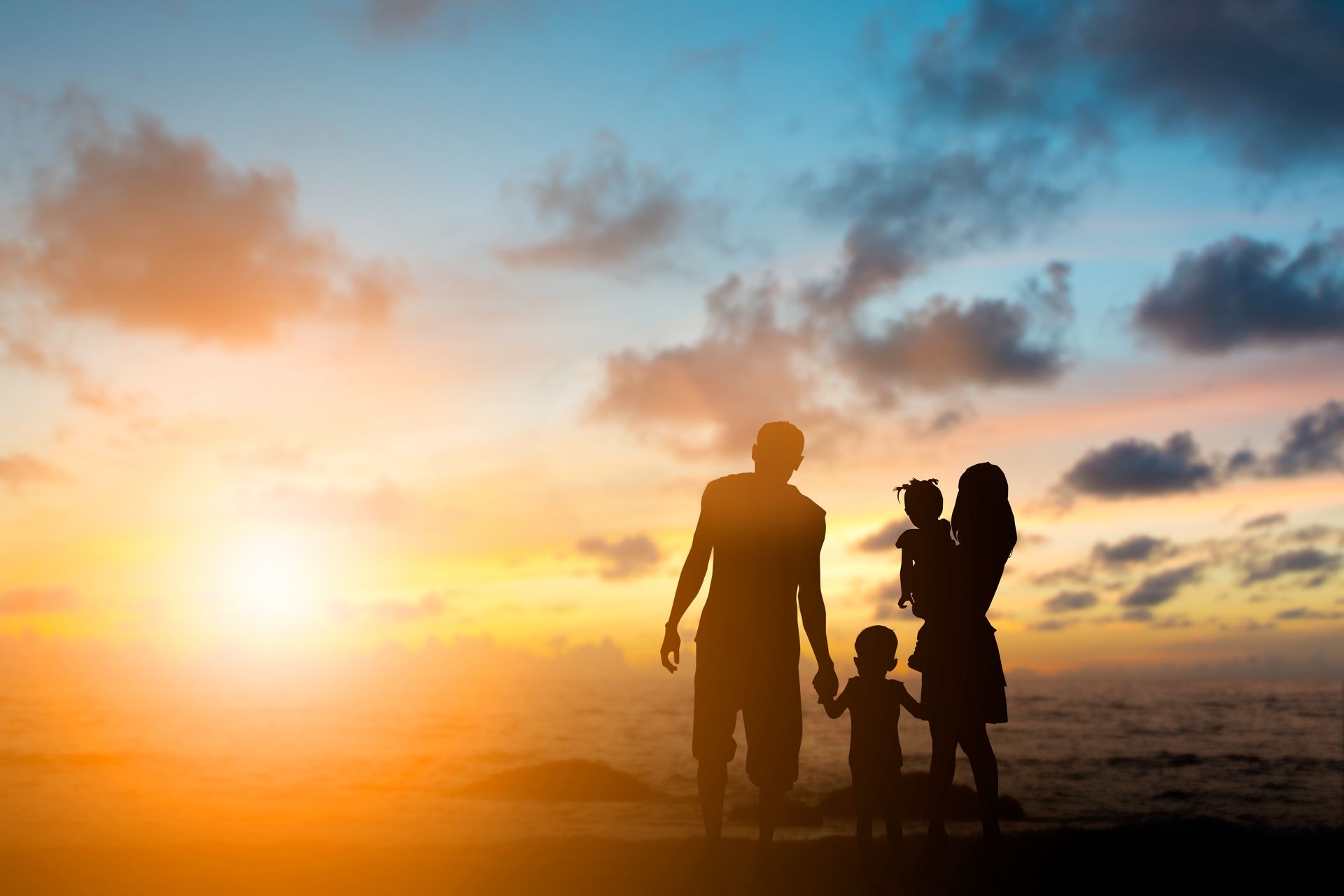 Silhouetted family of four standing on a beach at sunset, with warm orange sky and ocean waves. Silhouetted family of four standing on a beach at sunset, with warm orange sky and ocean waves.