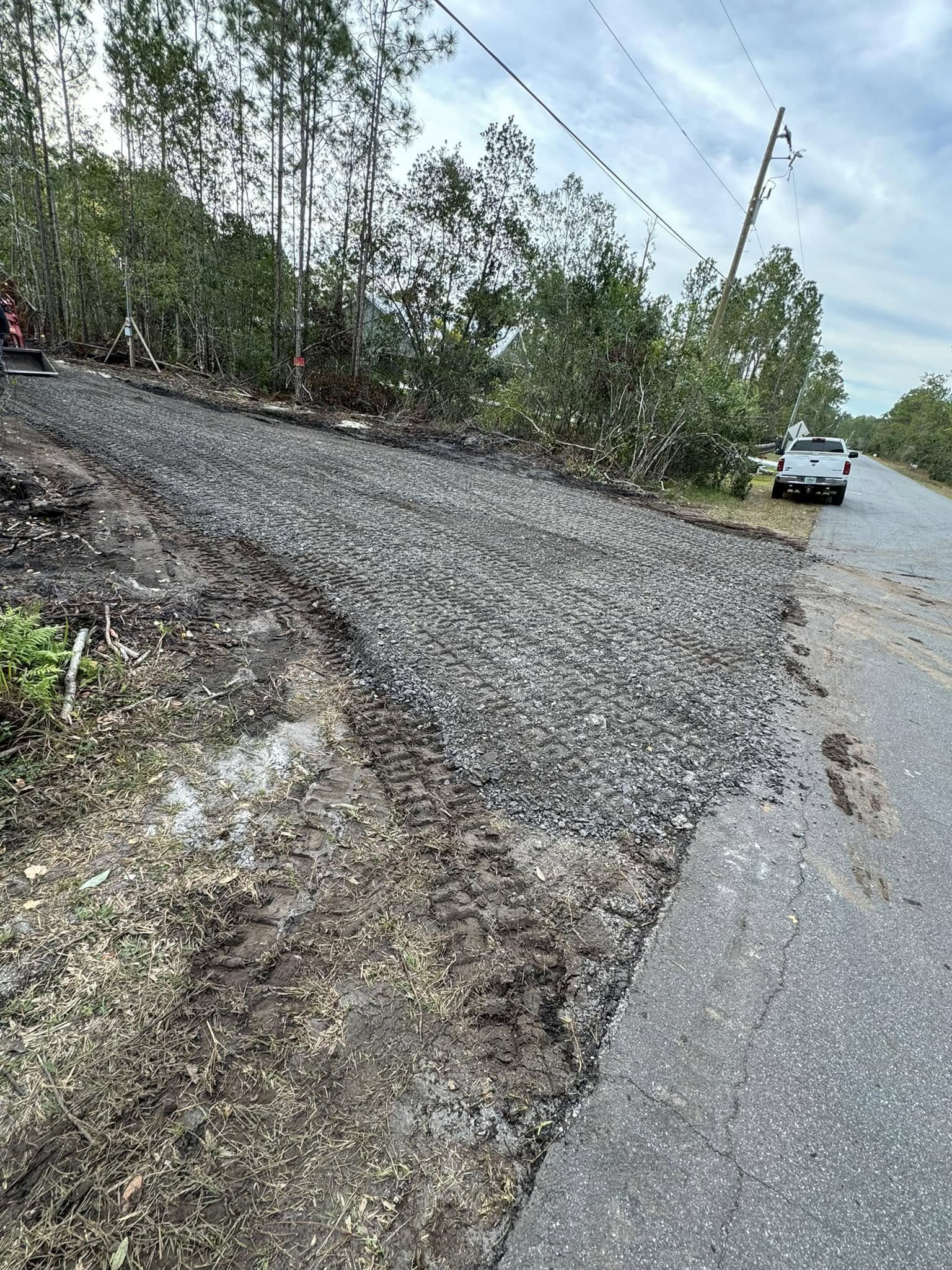 Roadside gravel slope next to paved road, power lines, and utility truck in a wooded area.