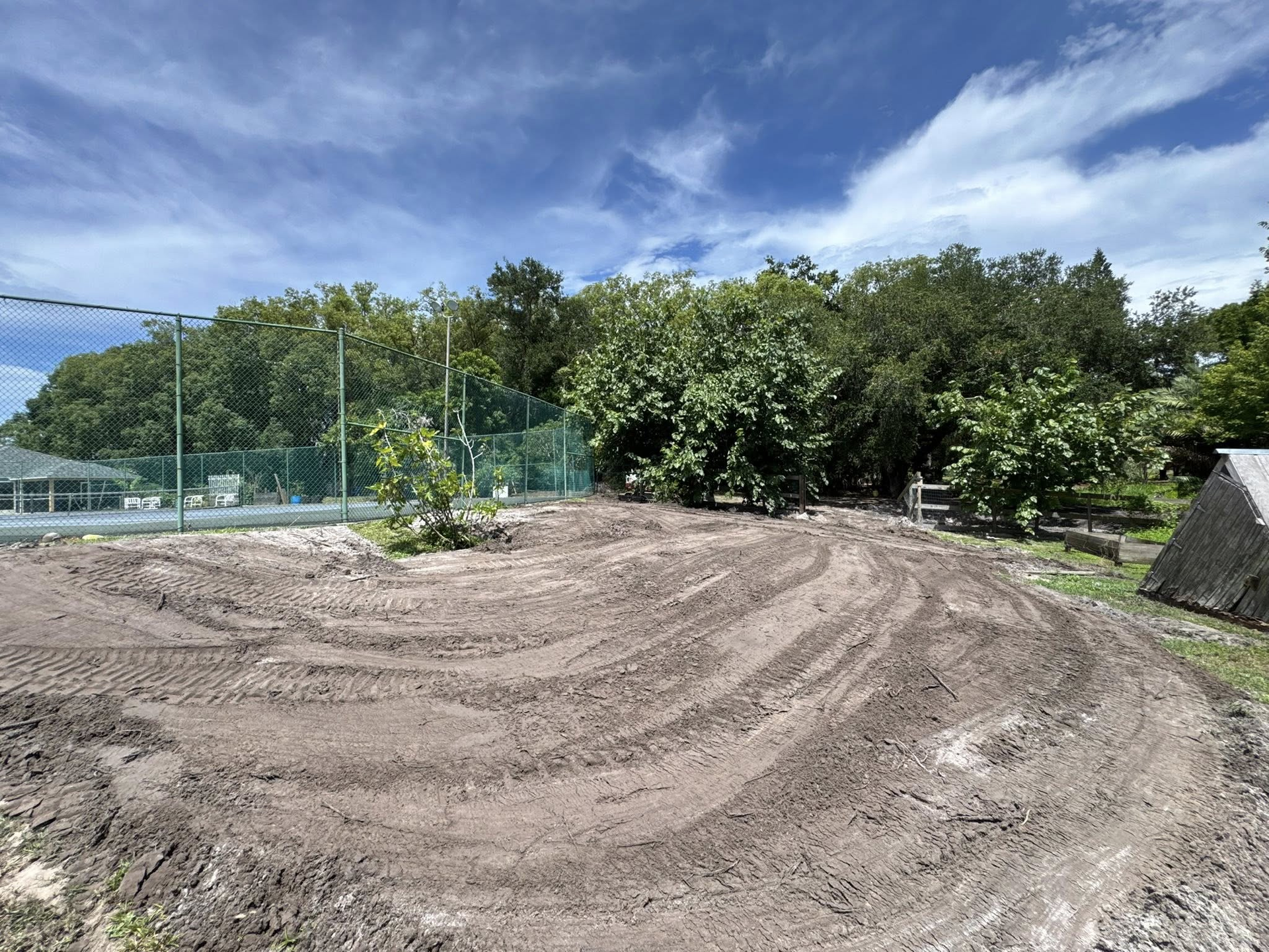 Dirt patch with tire tracks in front of trees and a tennis court, under a cloudy sky.