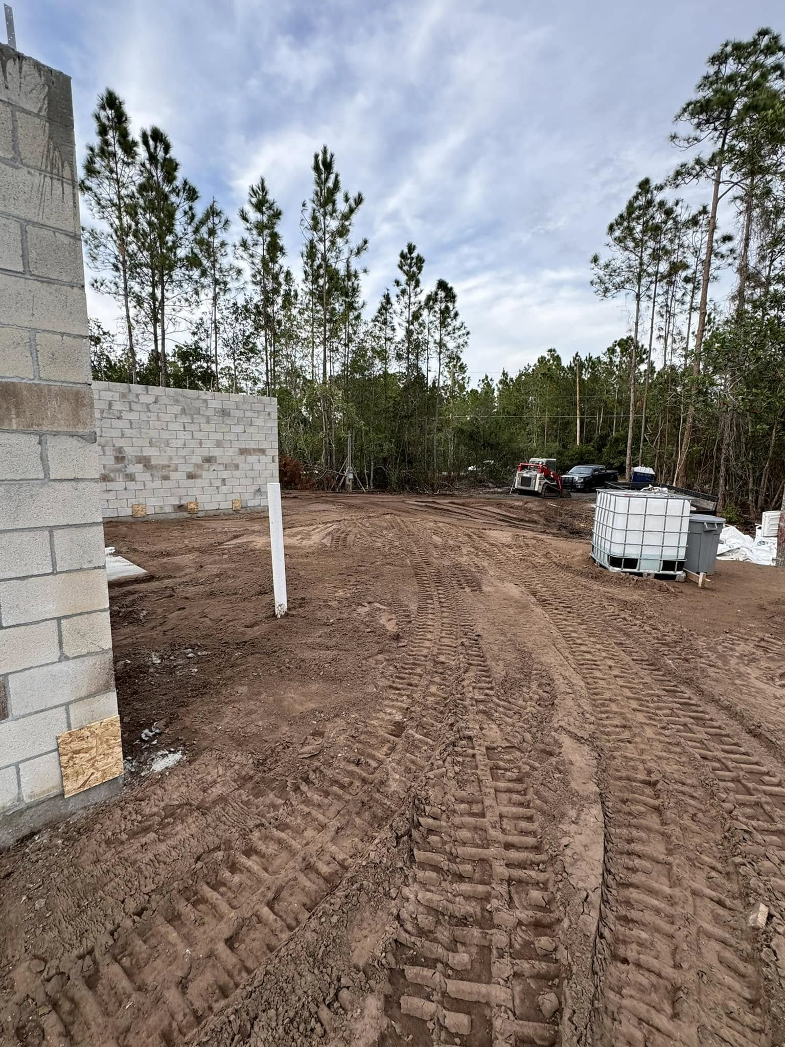 Construction site with concrete block walls, dirt, tire tracks, trees, and cloudy sky.