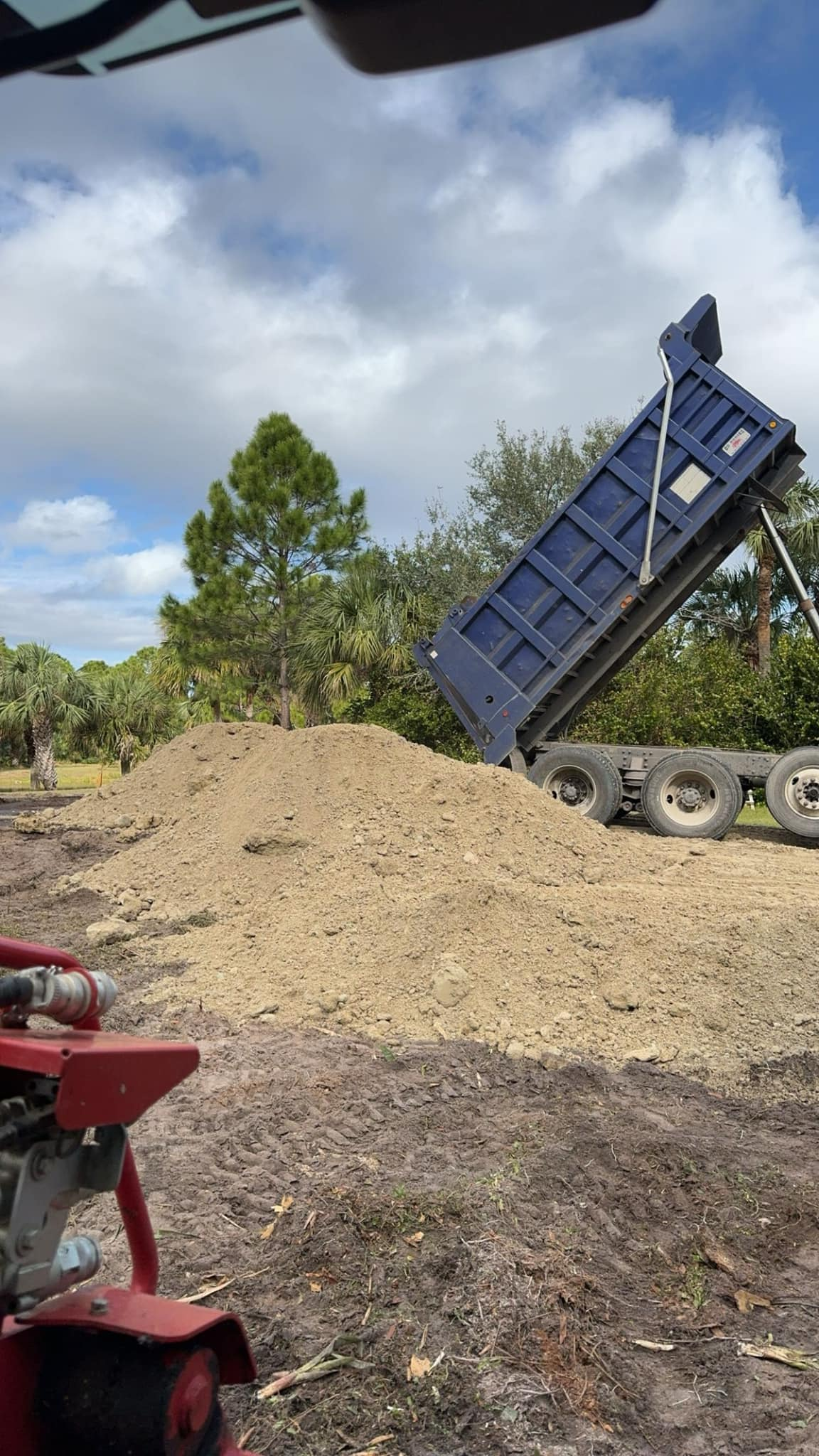 Dump truck dumping a load of dirt. Blue truck bed, tan dirt pile, cloudy sky.