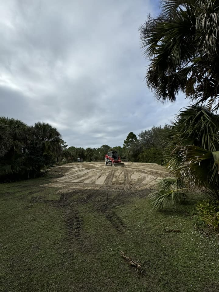 A red construction vehicle levels dirt in a grassy field under a cloudy sky, surrounded by trees.