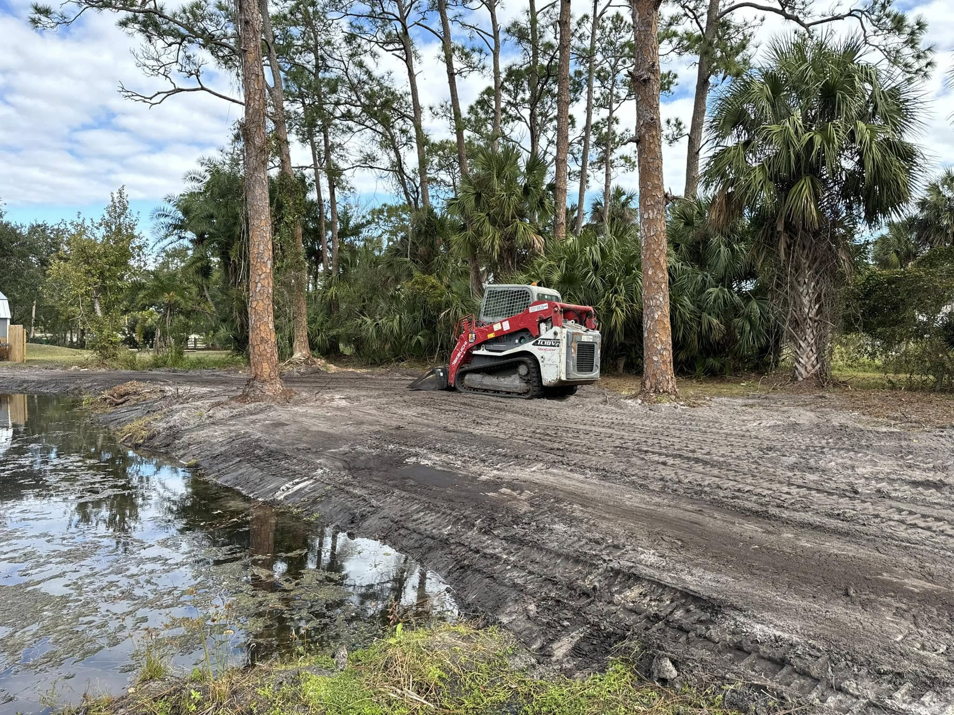 Skid steer loader on muddy ground near a body of water and trees.