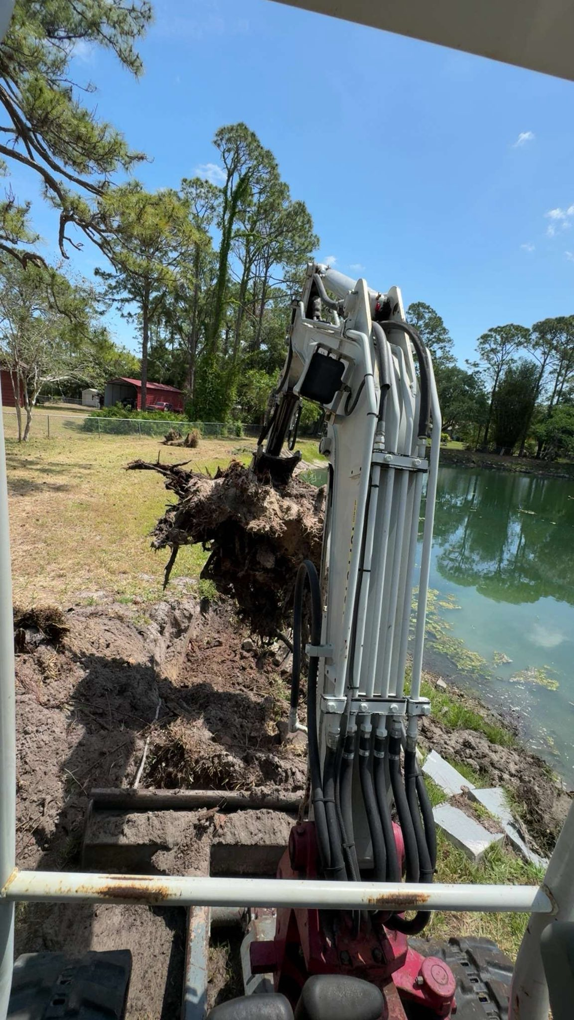 Mini excavator digging earth near a body of water with trees in the background under a blue sky.