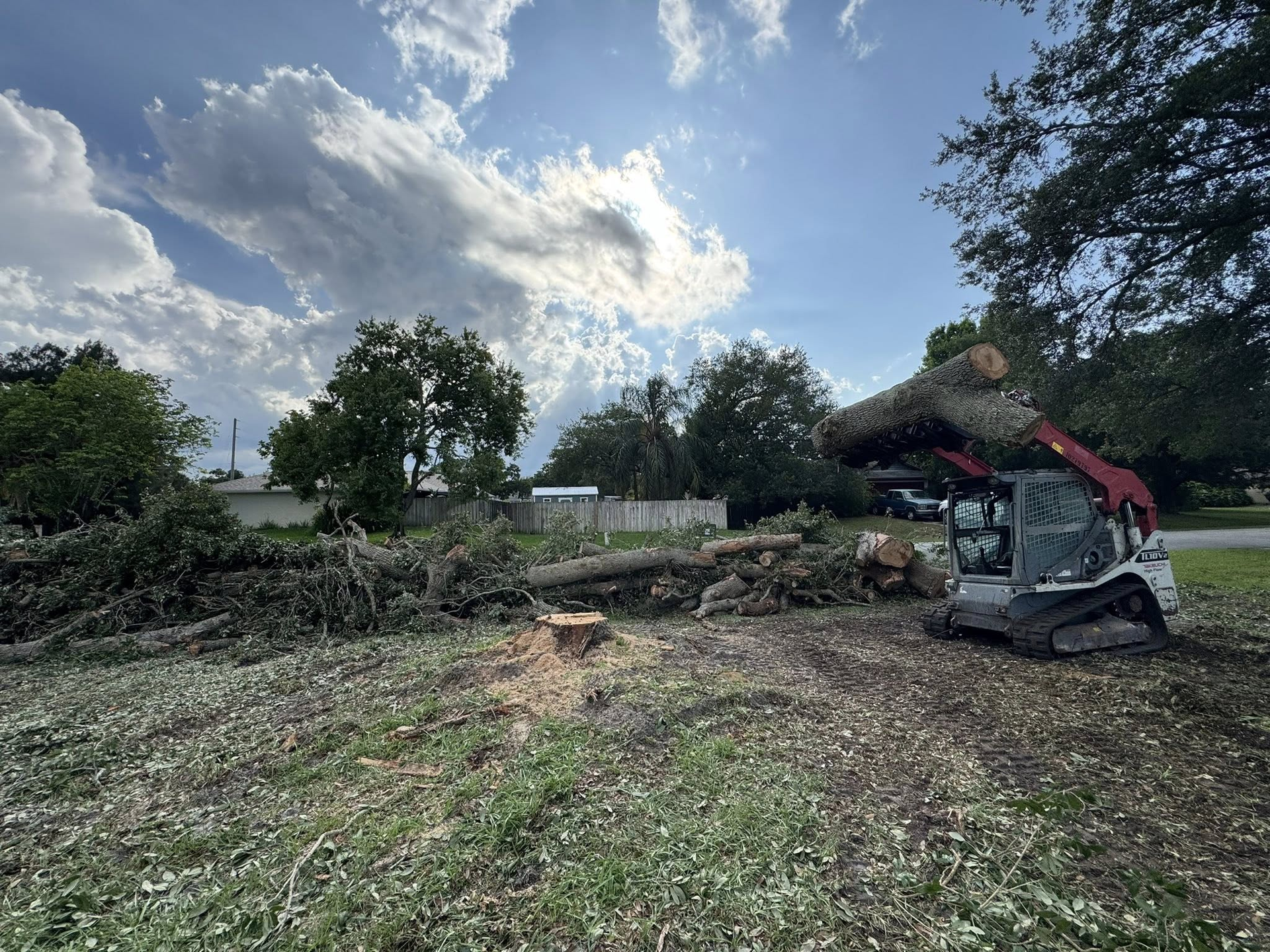 A Bobcat skid steer lifting a large tree branch over a debris pile on a grassy lot.