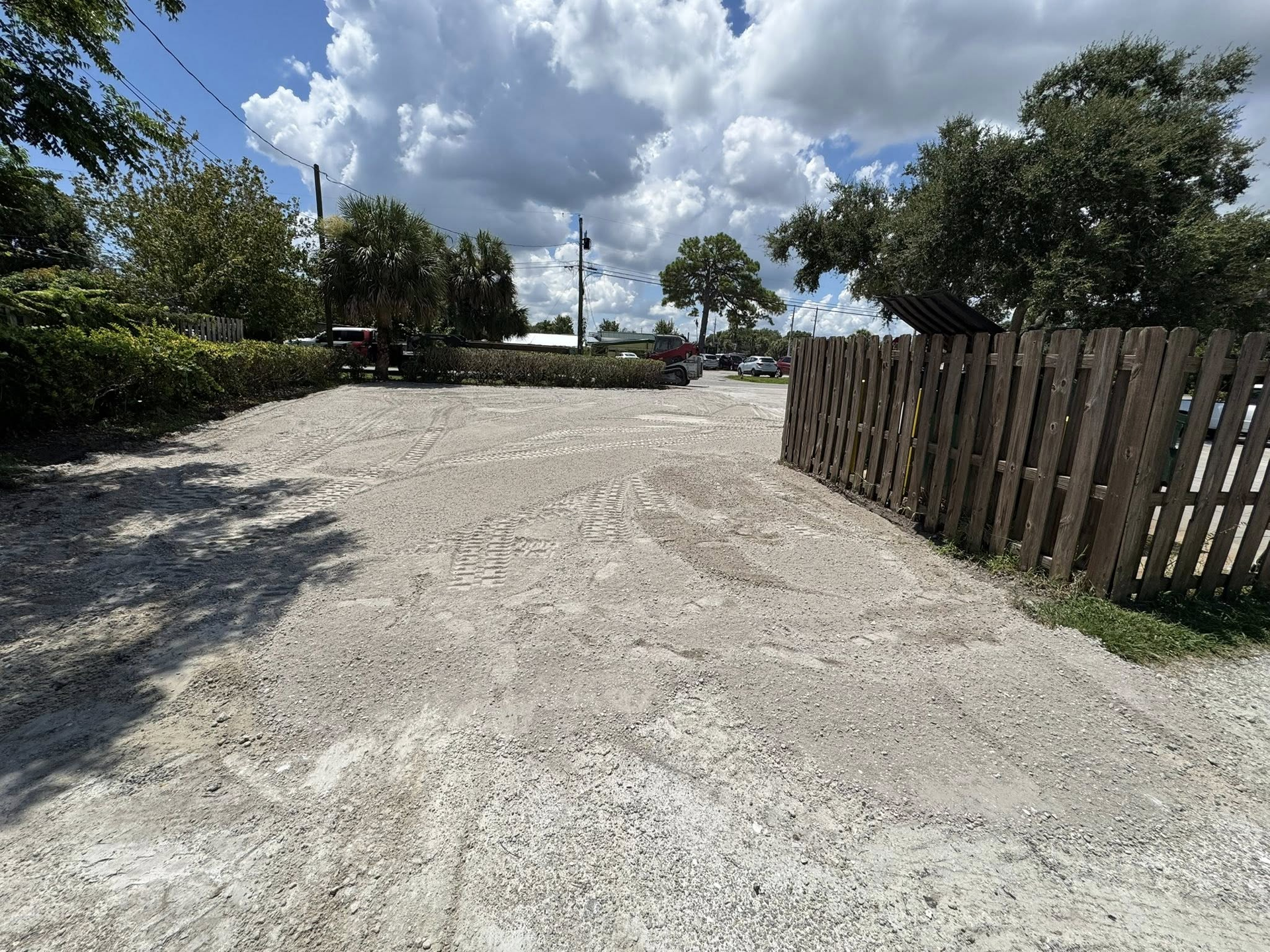 Empty gravel lot, wooden fence on right, trees and bushes surrounding, cloudy sky overhead.