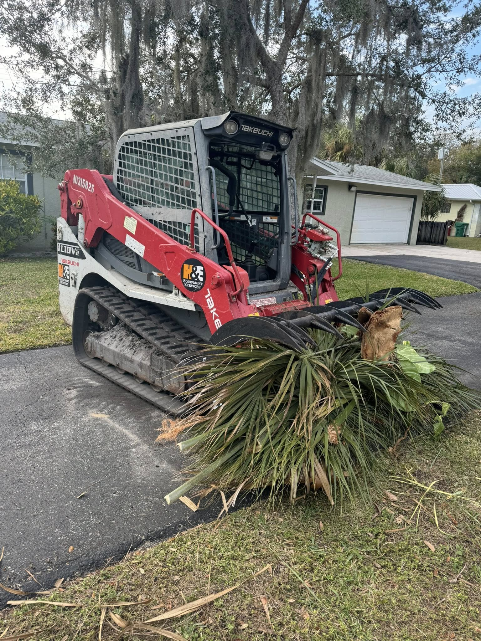 Red and white track skid steer with brush cutter attachment clearing debris on a driveway, houses in background.