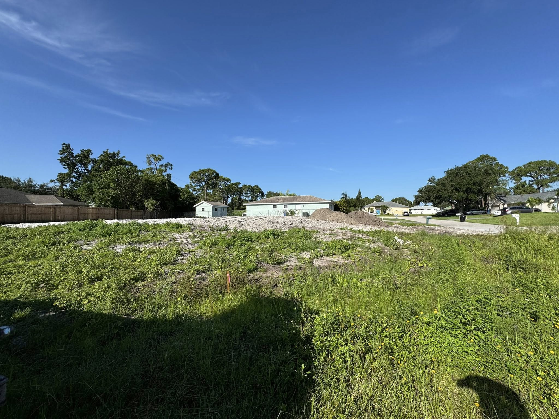 Grassy vacant lot with construction debris, houses in the distance, under a bright blue sky.