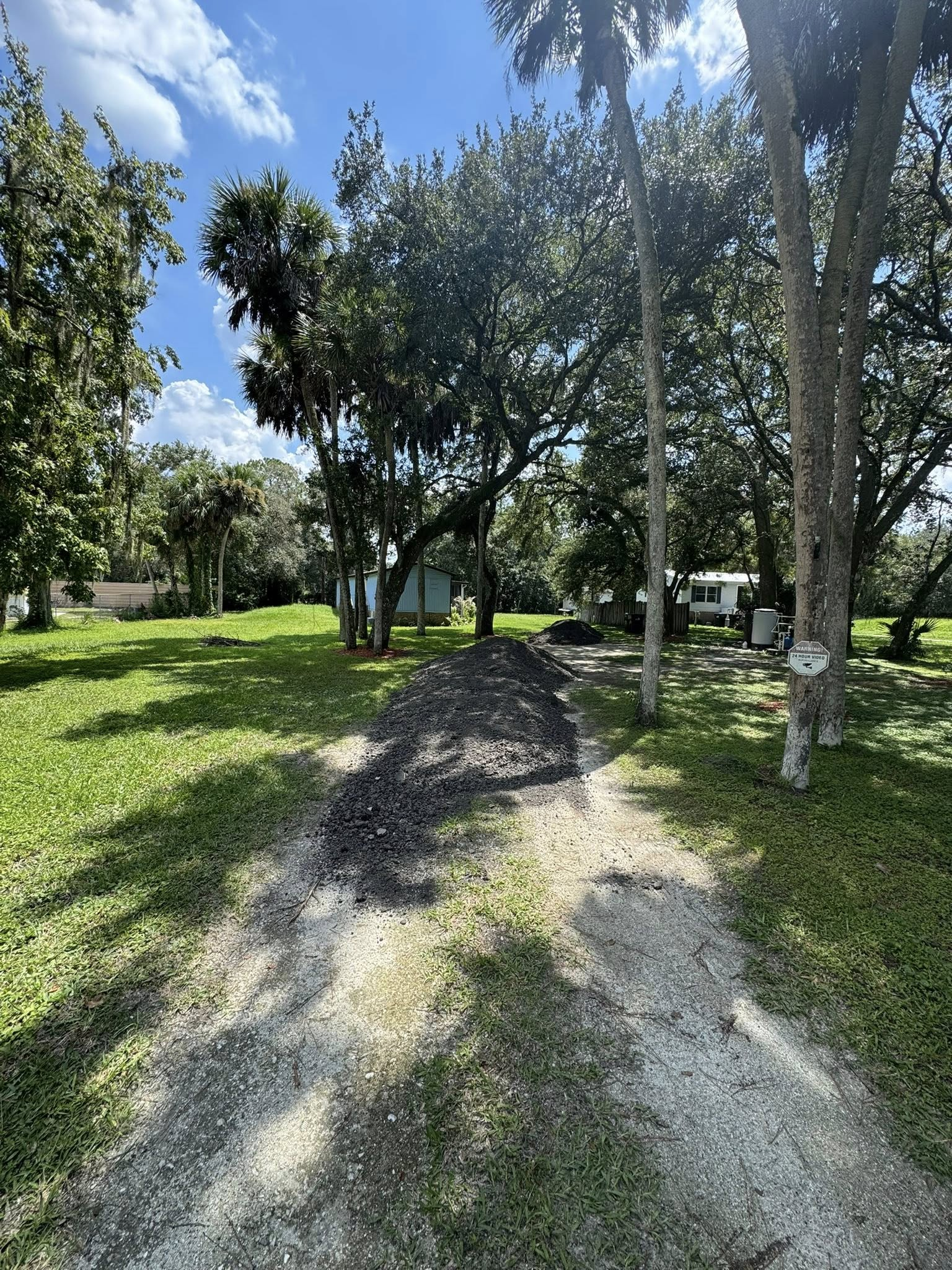 Dirt path leads through grassy yard to trees and small structures under a blue sky.