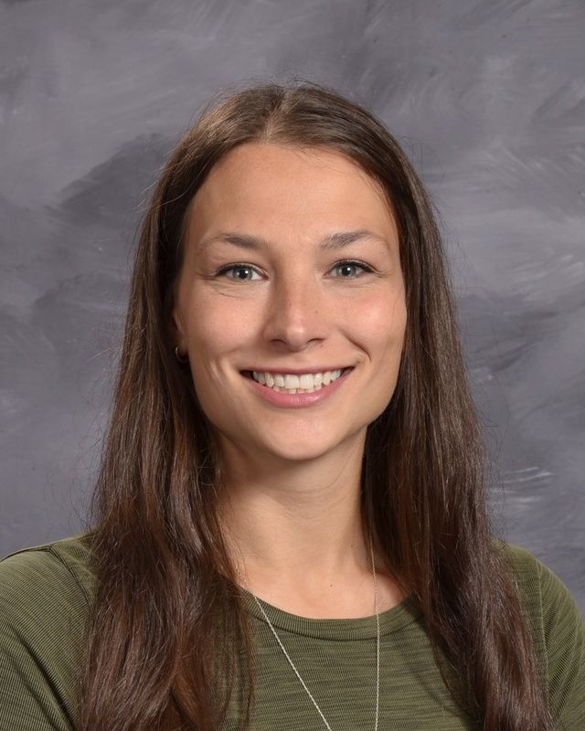 A woman with long brown hair is smiling and wearing a green shirt