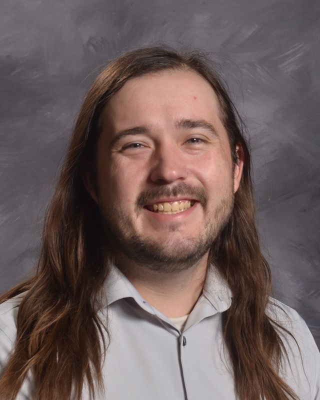 A man in a long hair and wearing a grey button up shirt smiling for the camera