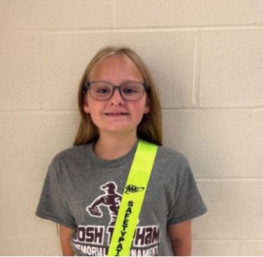 A young girl wearing a grey t-shirt and a safety patrol badge smiling for the camera
