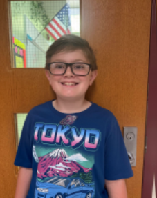 Young boy with light hair wearing glasses and a blue Tokyo t-shirt smiling for the camera