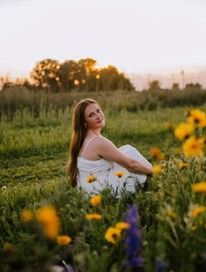 A young woman wearing a white dress sitting on the ground outside surrounded by flowers