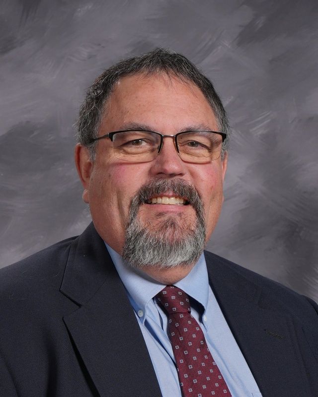 A man with a beard and glasses wearing a blue shirt smiling for the camera