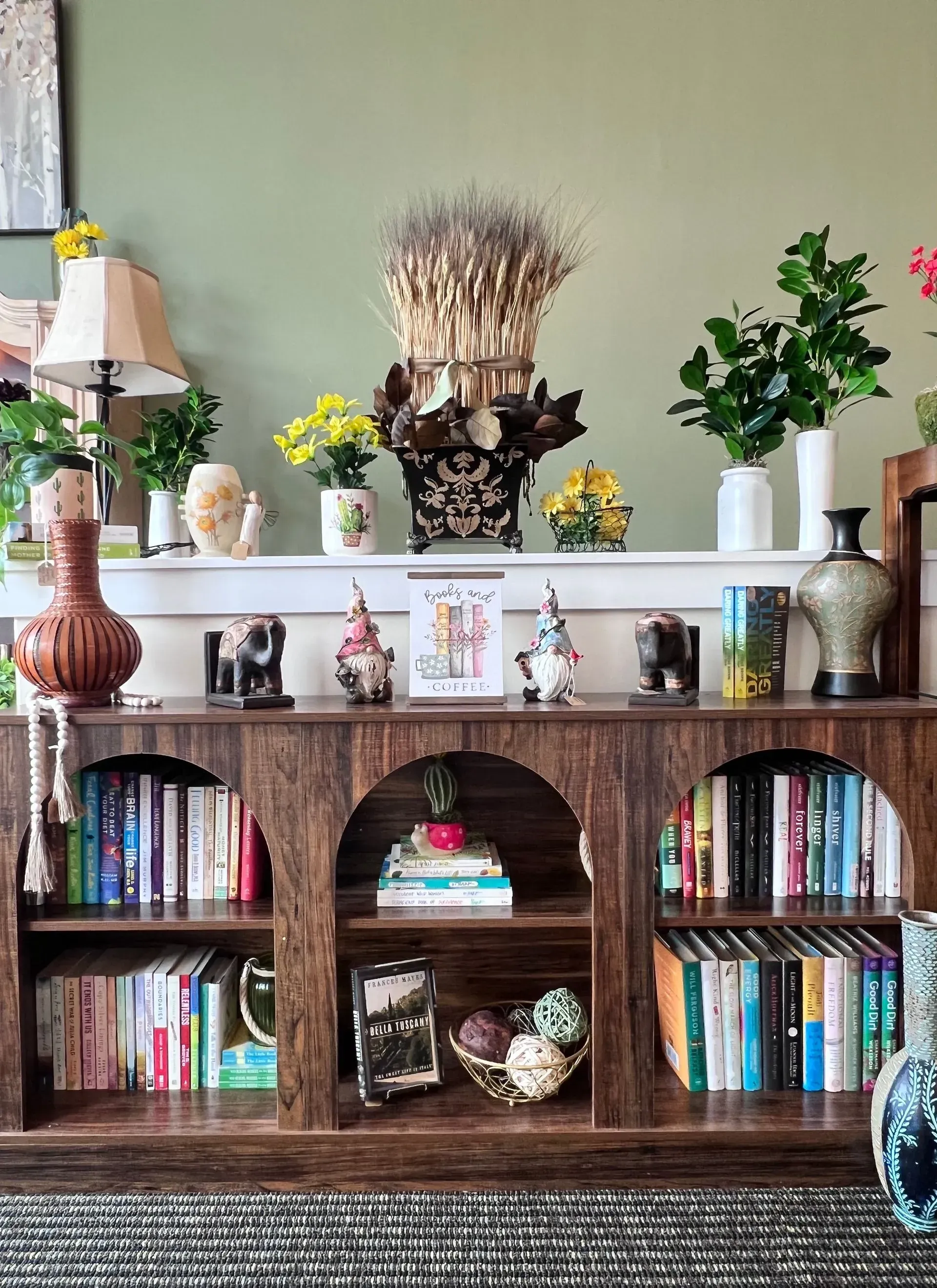 Wooden bookshelf with books and decor, topped with vases, flowers, and a lamp against a pale green wall