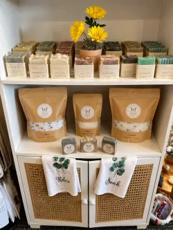 Shelf display of packaged bath products and soap bars with yellow flowers in a small planter