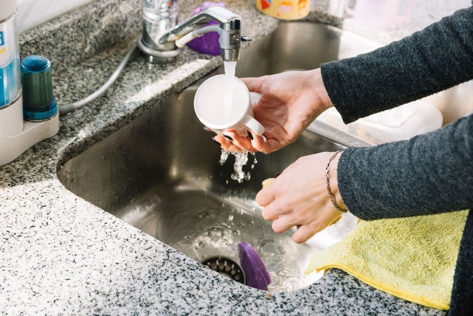 Hands rinsing a white cup over a kitchen sink with running water and a yellow towel nearby