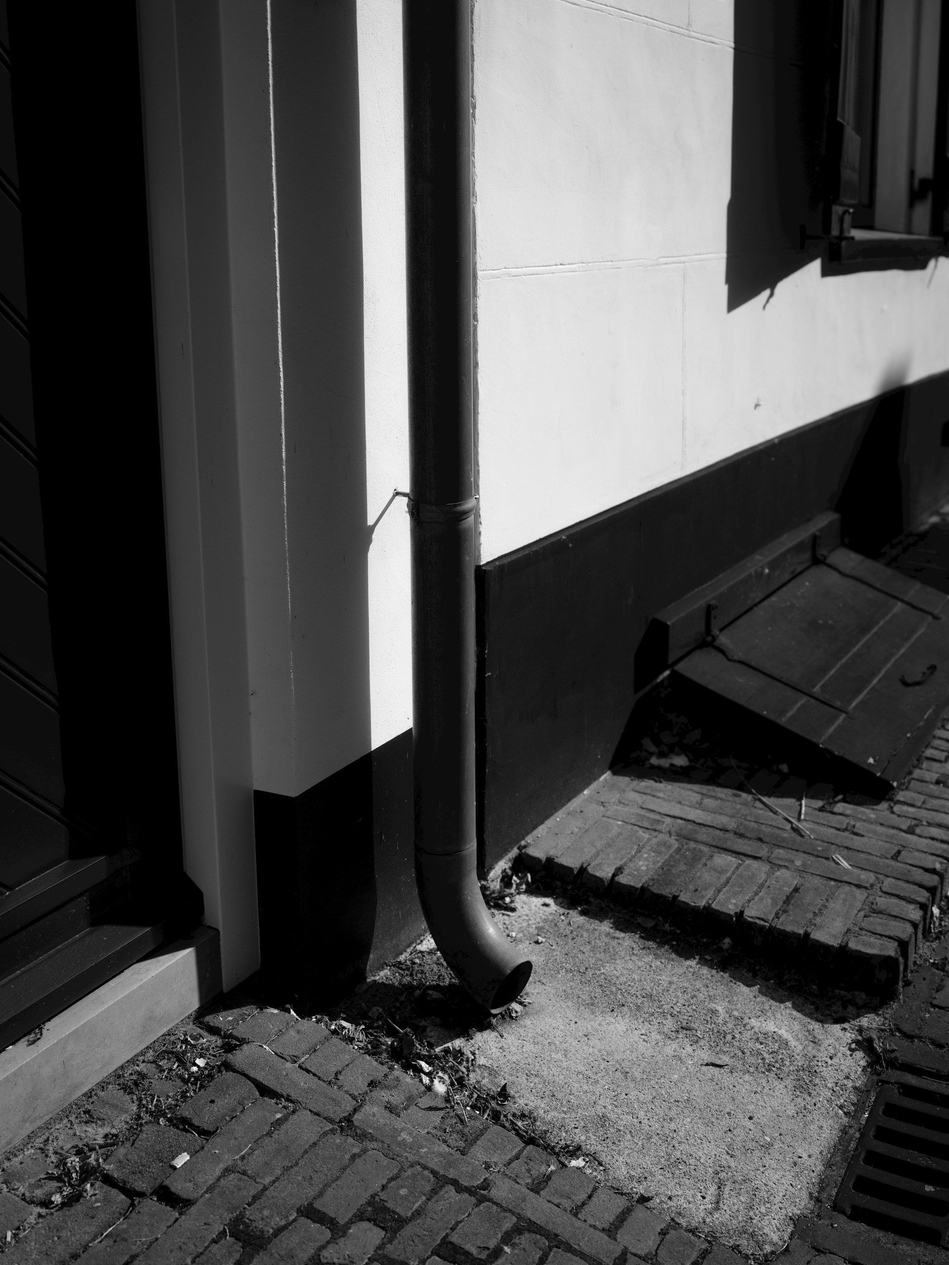 Black-and-white view of a building wall, doorway, and sidewalk with a small ramp and downspout.