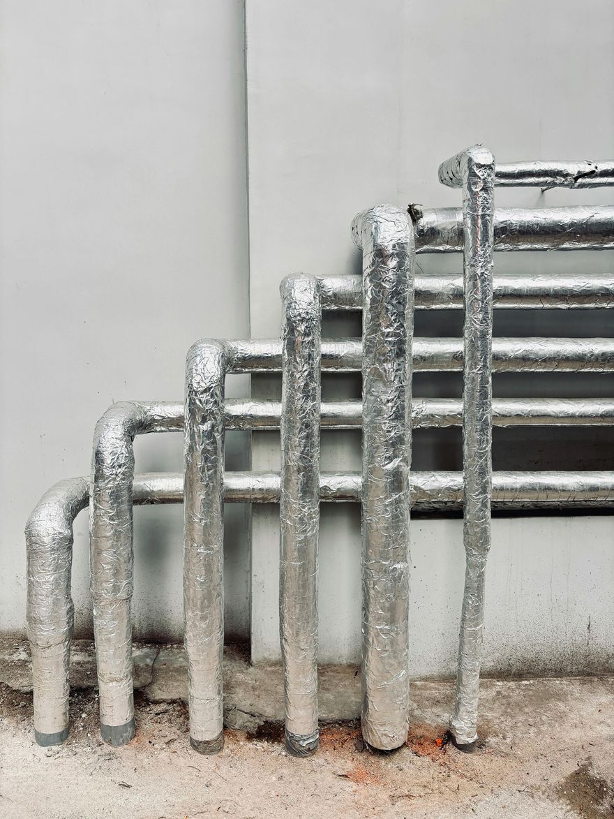Silver insulated pipes against a white wall, arranged in a row on a rough floor.