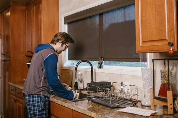 Person washing dishes at a kitchen sink beside a drying rack and wooden cabinets