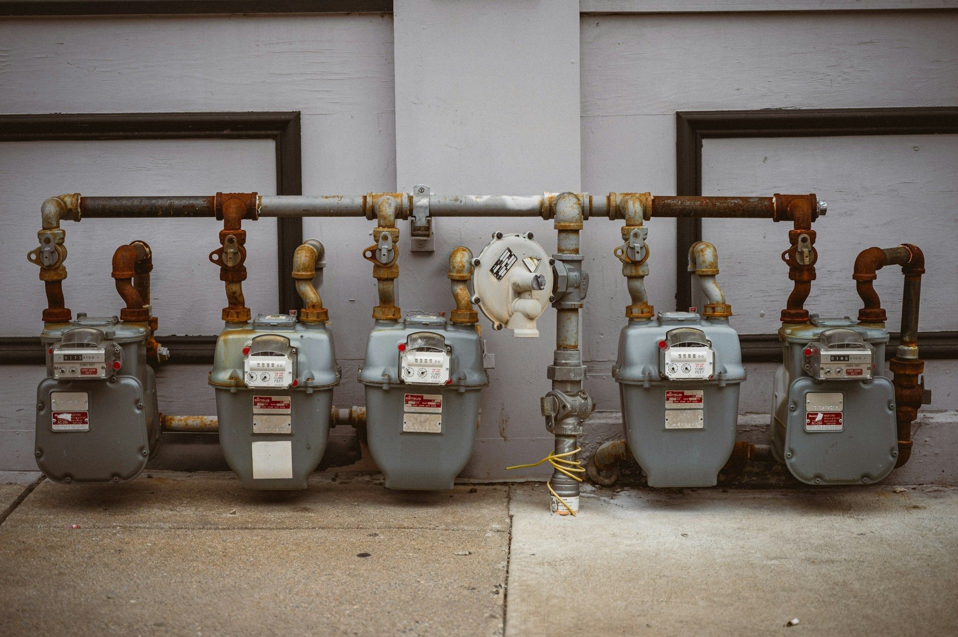 Five gas meters and pipes mounted on an exterior wall with concrete walkway below