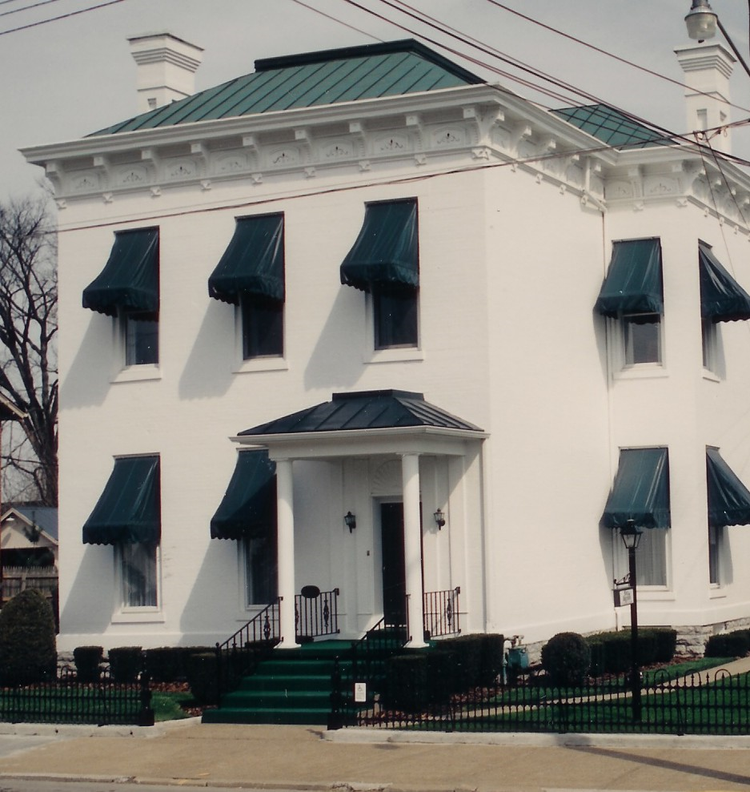 White two-story house with teal roof and window awnings, front porch, and black fence