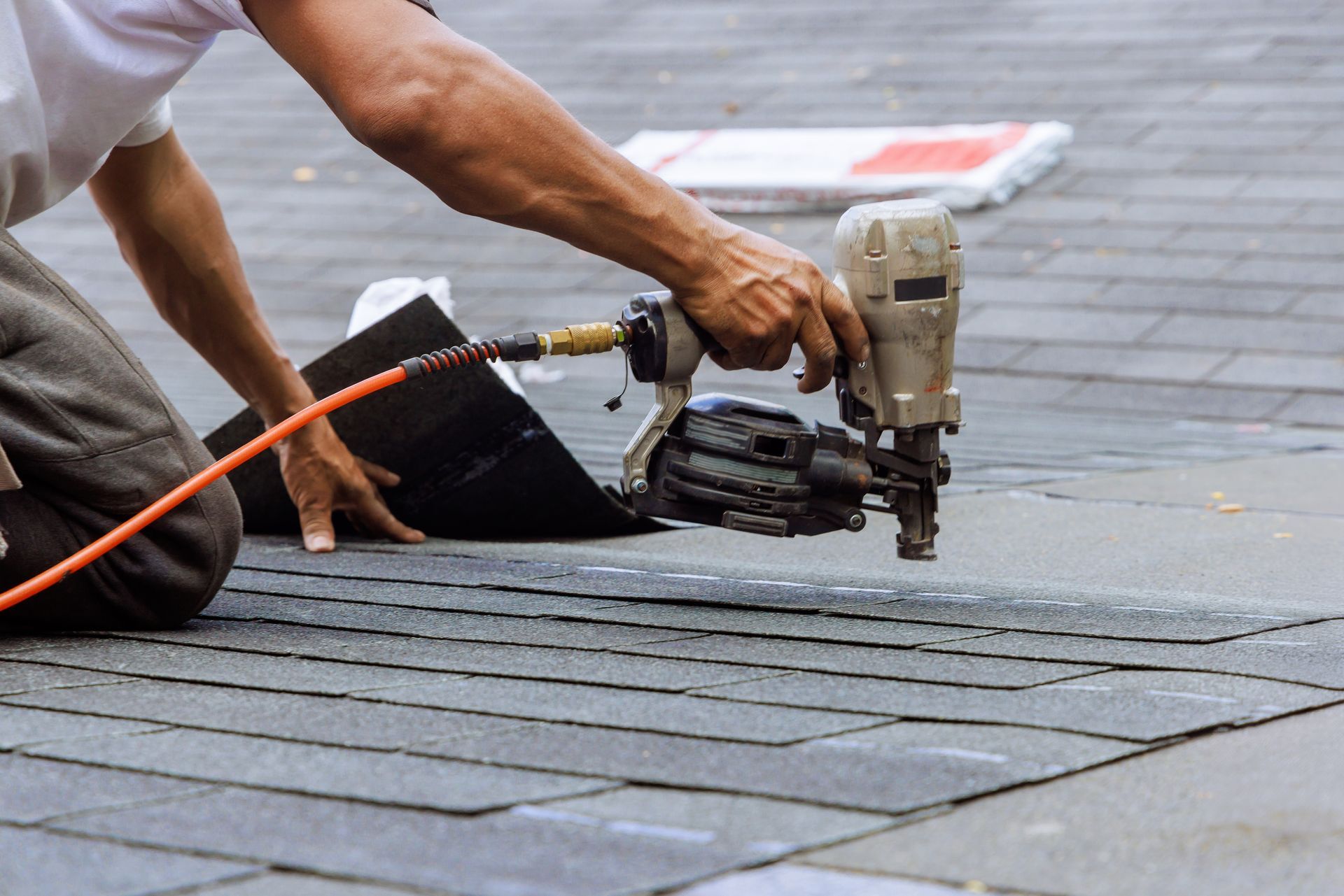 Roofer using a nail gun to attach shingles on a gray asphalt shingle roof.